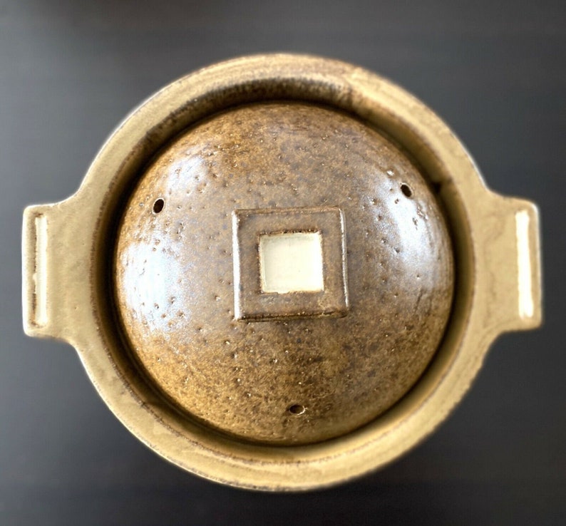 May include: A brown ceramic pot with a lid, featuring a textured surface and a small square opening in the center of the lid.