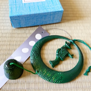 Peut inclure: Carillon en métal vert avec un motif de croissant de lune et une petite figurine d'oiseau. Le carillon est suspendu à une ficelle et a une petite cloche en bas.