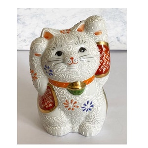 May include: White ceramic Maneki-Neko, or lucky cat, figurine with raised paw. The cat is decorated with intricate floral patterns in shades of blue, red, and gold. The cat is wearing a red and gold collar.
