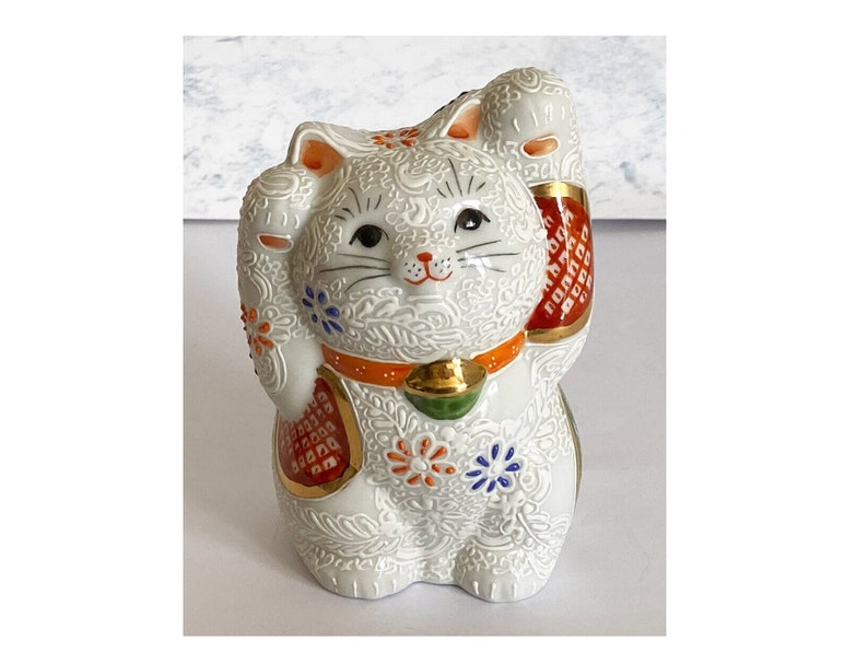May include: White ceramic Maneki-Neko, or lucky cat, figurine with raised paw. The cat is decorated with intricate floral patterns in shades of blue, red, and gold. The cat is wearing a red and gold collar.