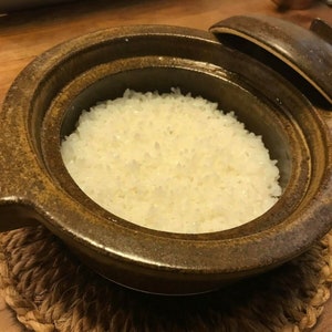 May include: A brown ceramic pot with a lid, filled with white cooked rice, sitting on a woven straw mat.