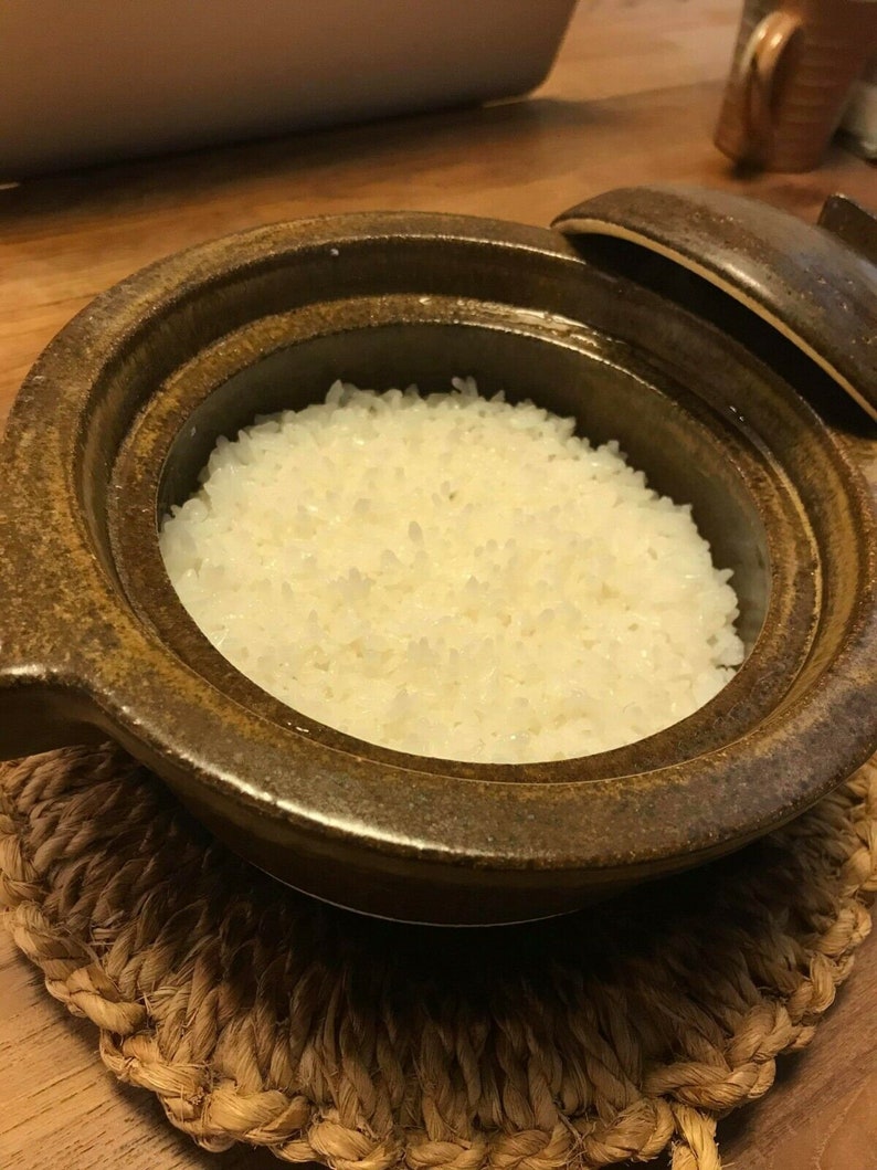 May include: A brown ceramic pot with a lid, filled with white cooked rice, sitting on a woven straw mat.