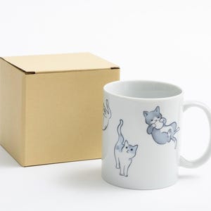 以下が含まれることがあります： 白のセラミックマグカップ。グレーと白の遊び心のある猫のイラストが描かれています。マグカップの隣には、茶色の段ボール箱があります。 マグカップにはカーブしたハンドルがあり、白い背景に置かれています。