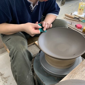 May include: A person uses a green sanding tool to smooth the edge of a brown ceramic bowl on a pottery wheel. The bowl is on a wooden base and the wheel is on a metal base.