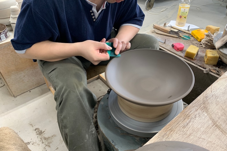 May include: A person uses a green sanding tool to smooth the edge of a brown ceramic bowl on a pottery wheel. The bowl is on a wooden base and the wheel is on a metal base.
