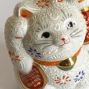 May include: White ceramic Maneki-Neko, or lucky cat, figurine with raised paw. The cat is decorated with intricate floral patterns in shades of orange, blue, and gold. The cat has black eyes and a red nose.