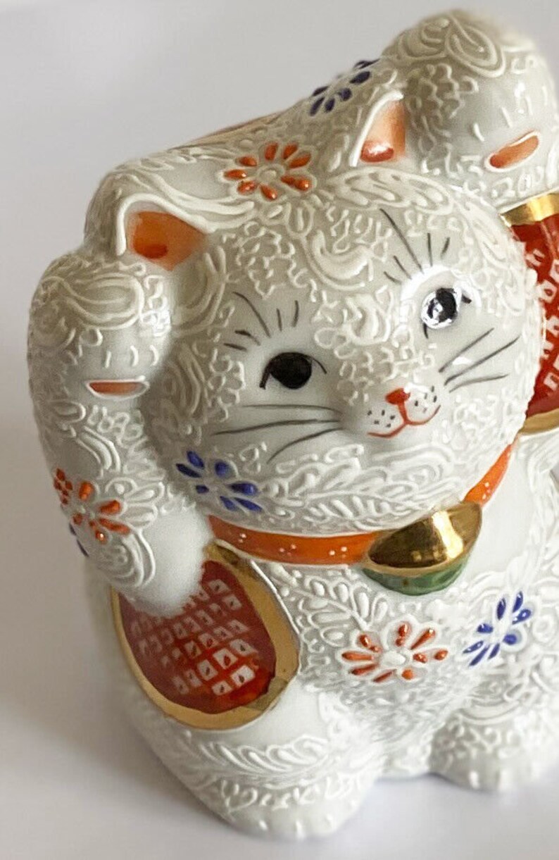 May include: White ceramic Maneki-Neko, or lucky cat, figurine with raised paw. The cat is decorated with intricate floral patterns in shades of orange, blue, and gold. The cat has black eyes and a red nose.