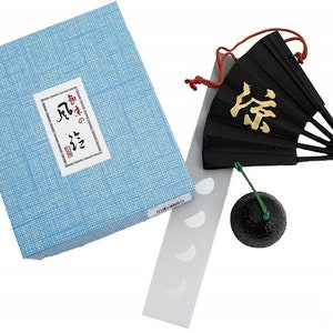 Peut inclure: Un carillon japonais noir et or avec un charme en forme d'éventail et une cloche verte et noire. Le carillon est suspendu à une ficelle rouge et a un fond blanc avec une boîte à motifs bleus et blancs.
