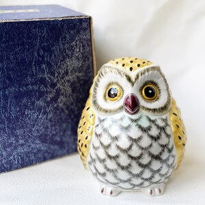 May include: A ceramic owl figurine with yellow, white, and black markings. The owl has large, round eyes with black pupils and gold accents. The owl is sitting on a white surface.