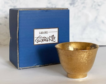 Könnte beinhalten: Eine goldfarbene Sake-Schale aus Keramik mit gehämmerter Textur. Die Schale steht auf einer weißen Oberfläche neben einer blauen Box mit japanischem Text, der "伝統と創造" (Tradition und Kreation) lautet.