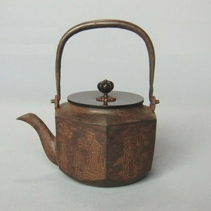 May include: A brown cast iron teapot with a black lid and a black knob. The teapot has a decorative design of figures on the sides.