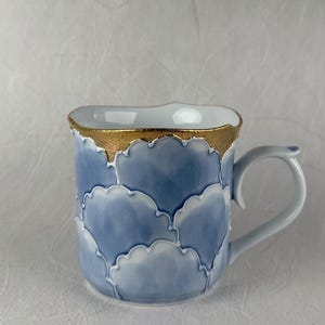 May include: A ceramic mug featuring a repeating blue and white scalloped pattern across the exterior. The top rim of the mug is finished with a textured gold metallic band. The mug has a curved handle on the right side and a white interior.