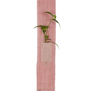 May include: A pink fabric wall hanging with a small pocket that holds a green plant. The hanging has tassels at the bottom.