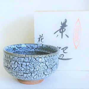 Puede incluir: Un cuenco de té de cerámica con un esmalte craquelado azul claro y blanco. El cuenco se asienta sobre una base marrón. Detrás del cuenco hay una caja blanca con caligrafía japonesa.