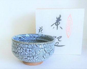May include: A ceramic tea bowl with a light blue and white crackle glaze. The bowl sits on a brown base. A white box with Japanese calligraphy is behind the bowl.