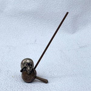 May include: A silver metal incense holder with a small animal figure on top. The holder is attached to a long, thin, brown incense stick.