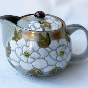 以下が含まれることがあります： グレーのベースと蓋が付いた小さなセラミックティーポット。白い花の模様と緑の葉で飾られています。ティーポットは茶色のハンドルと注ぎ口、蓋には小さな茶色のノブが付いています。花の模様には黄色の中心が含まれています。