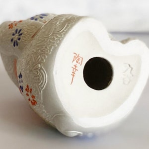 May include: White ceramic figurine of a seated woman with floral details in blue, red, and orange. The figurine has a hole in the bottom and is marked with Japanese characters.