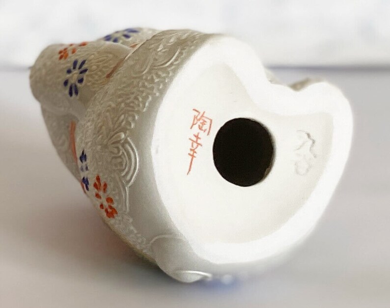 May include: White ceramic figurine of a seated woman with floral details in blue, red, and orange. The figurine has a hole in the bottom and is marked with Japanese characters.