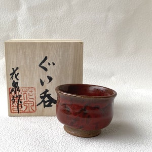 May include: A small, red ceramic teacup with a brown rim sits on a white surface. The teacup is in a wooden box with Japanese characters written on the side. The characters read "ぐり茶" which translates to "green tea".