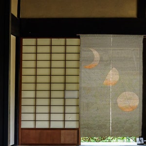 Pode incluir: Interior japonês tradicional com uma tela Shoji e uma cortina suspensa. A cortina apresenta três desenhos de fases lunares em laranja e branco. A tela é uma grade de molduras de madeira e papel translúcido.