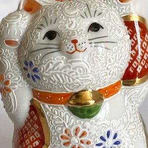May include: White ceramic Maneki-Neko, or lucky cat, figurine with raised paw, orange and gold accents, and intricate floral patterns.