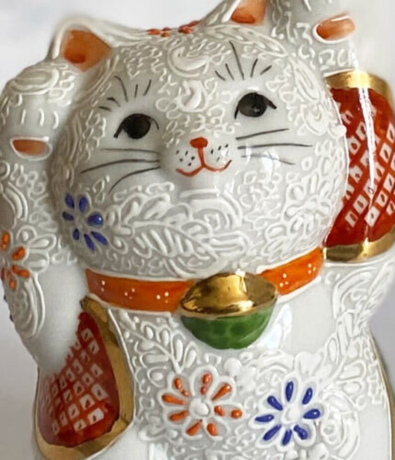 May include: White ceramic Maneki-Neko, or lucky cat, figurine with raised paw, orange and gold accents, and intricate floral patterns.