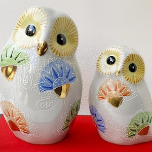 May include: Two ceramic owl figurines. The owls are light gray with intricate floral designs in orange, green, and blue. They have large yellow eyes with black pupils and gold accents on the beak and wings. The owls are sitting on a red surface.