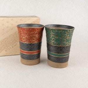 May include: Two ceramic tumblers with a textured finish. One features a red upper section with gold vine patterns, while the other has a green upper section with gold vines. Both have a black middle band with a coloured stripe and a tan base.