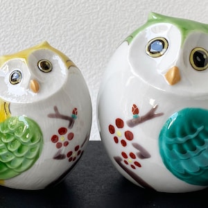 May include: Two ceramic owl figurines. One owl is yellow and white with green accents, the other is green and white with teal accents. Both have brown branches with red berries and are decorative.