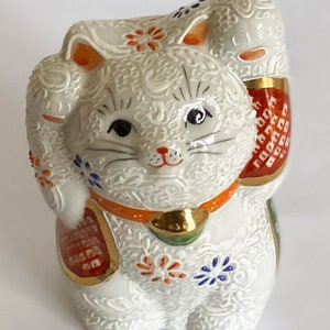 May include: White ceramic Maneki-Neko, or lucky cat, figurine with raised paw, gold accents, and intricate floral patterns. The cat has black eyes and a red collar.