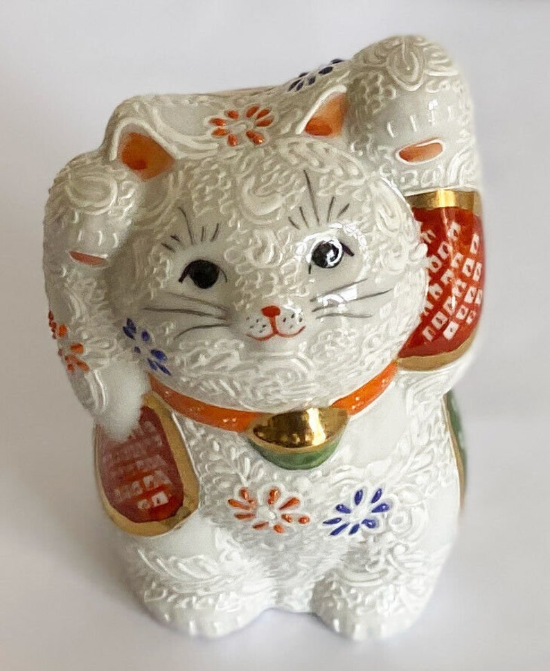 May include: White ceramic Maneki-Neko, or lucky cat, figurine with raised paw, gold accents, and intricate floral patterns. The cat has black eyes and a red collar.