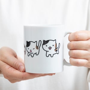May include: White ceramic mug featuring three cartoon cats with black, brown, and white markings. The cats have different expressions, and the mug is held by a person. The mug has a white handle.