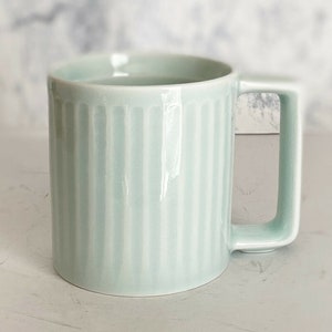 Op de afbeelding: Een lichtblauwe keramische mok met verticale ribbels. De mok heeft een handvat en is perfect voor het genieten van een warme drank.