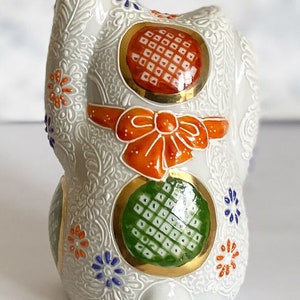 May include: White ceramic Maneki-Neko, or lucky cat, figurine with orange, green, and gold accents. The cat is decorated with a bow and intricate floral patterns.