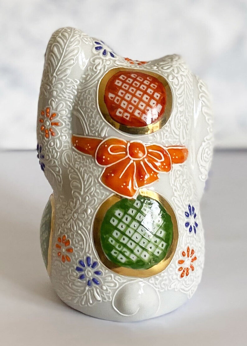 May include: White ceramic Maneki-Neko, or lucky cat, figurine with orange, green, and gold accents. The cat is decorated with a bow and intricate floral patterns.