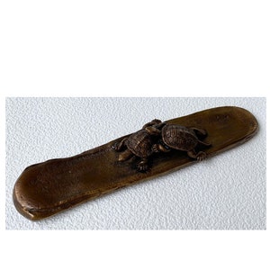 May include: A long, flat, brown, metal incense burner with two turtles on top. The turtles are facing each other and are made of the same metal as the burner.