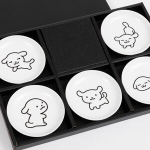 以下が含まれることがあります： 犬が様々なポーズで描かれた黒い輪郭の白いセラミック製の皿6枚セット。皿は蓋付きの黒い箱に並べられています。