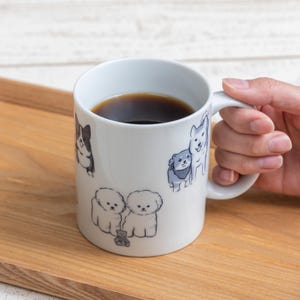 以下が含まれることがあります： 様々な犬種の青とグレーのイラストが描かれた、濃い液体が入った白いセラミックマグカップ。 マグカップは手で持たれており、木製のトレイの上に置かれています。 マグカップの高さは約10cmです。