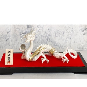 May include: A white dragon figurine with gold accents holding a clear glass orb. The dragon is sitting on a red velvet base with a black wooden frame. The base has a small wooden tag with the text "同型福" in Japanese.