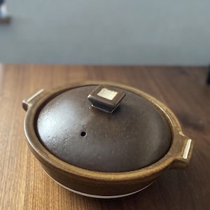 May include: A brown ceramic pot with a lid and two handles. The lid has a small square knob on top.