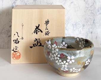 Könnte beinhalten: Eine Teeschale aus Keramik mit hellblauer Glasur und einem Muster aus weißen Kirschblüten. Die Schale befindet sich in einer Holzkiste mit japanischen Schriftzeichen, die "Teeschale" bedeuten.