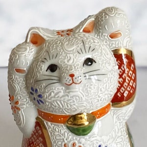 May include: White ceramic Maneki-Neko, or lucky cat, figurine with raised paw, gold bell, and intricate orange, red, and blue floral patterns.