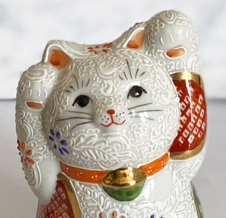 May include: White ceramic Maneki-Neko, or lucky cat, figurine with raised paw, gold bell, and intricate orange, red, and blue floral patterns.