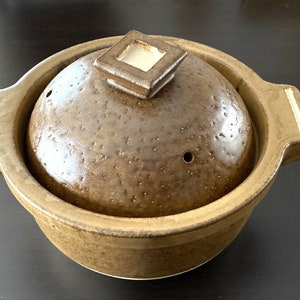 May include: A brown ceramic pot with a lid. The pot has a speckled brown glaze and a small hole in the lid. The lid has a square design with a raised edge.