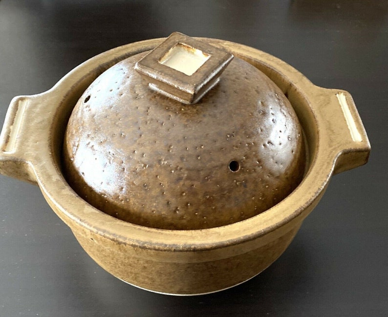 May include: A brown ceramic pot with a lid. The pot has a speckled brown glaze and a small hole in the lid. The lid has a square design with a raised edge.