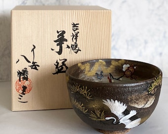 以下が含まれることがあります： 手描きの白い鶴、金色の松葉、赤い梅の花が描かれた濃い茶色の陶器製の茶碗。茶碗には、黒い日本語の書道と赤い印章が施された明るい色の木製収納箱が付属しています。