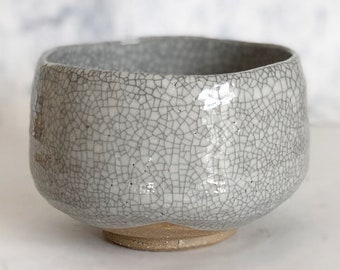 May include: A gray ceramic bowl with a crackle glaze finish. The bowl has a rounded shape with a wide opening and sits on a small, unglazed base. The surface is covered in a network of fine lines.