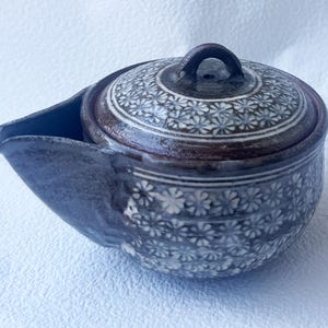 May include: A ceramic teapot with a lid, featuring a dark brown rim and handle. The body and lid are decorated with a repeating floral pattern in white and blue. The spout is integrated into the design.