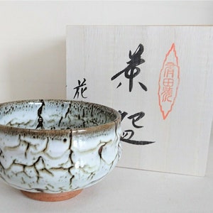 May include: A white ceramic tea bowl with a brown rim and a crackled glaze. The bowl is sitting on a wooden box with Japanese characters written on it.